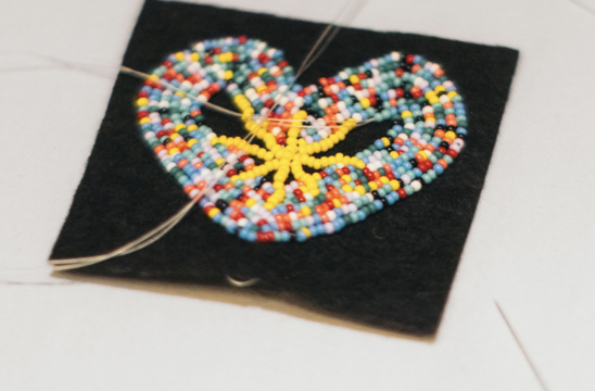Beaded heart pendant stitched with colorful seed beads on a black fabric square, with thread trailing from the edges