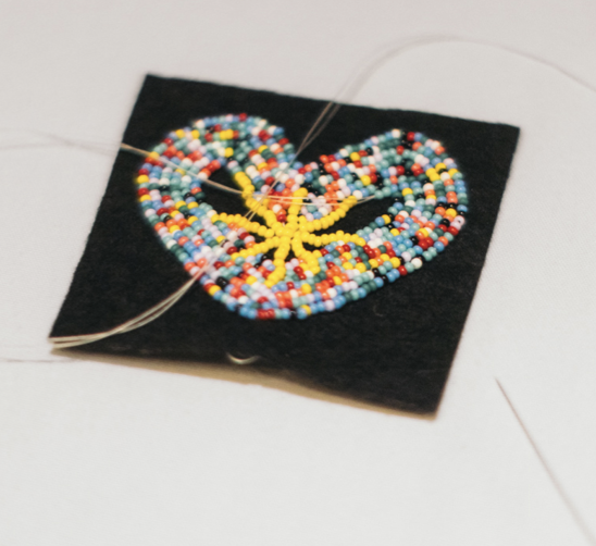 Beaded heart pendant stitched with colorful seed beads on a black fabric square, with thread trailing from the edges