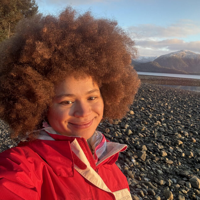 Selfie of a person with large curly hair in a bright red jacket at a rocky shoreline with mountains in the distance.