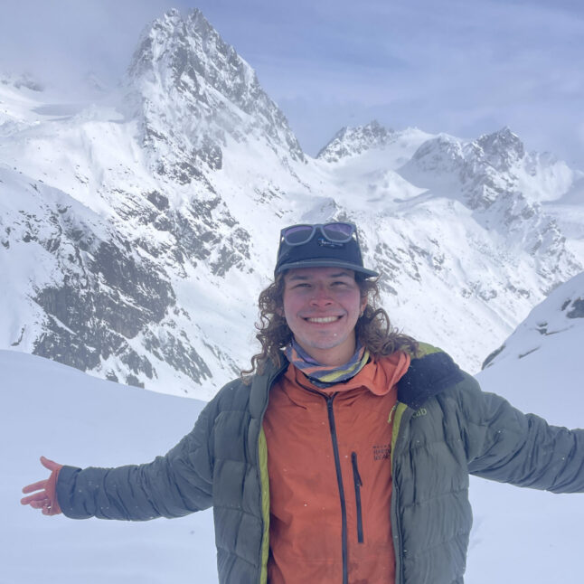 Smiling hiker in orange jacket and green shell layers stands in deep snow with mountains behind, arms outstretched.