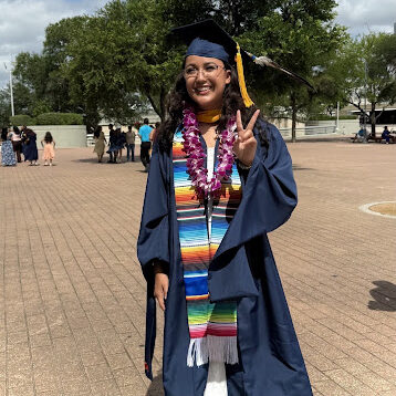Graduate in a navy cap and gown posing with a peace sign, wearing a colorful stole and purple lei; towers and trees in the background on a paved plaza.]